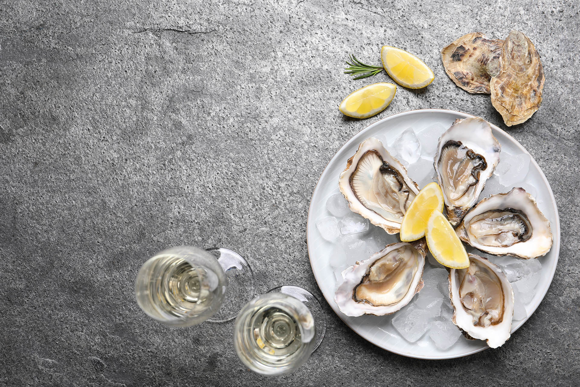 Fresh oysters with lemon, rosemary and glasses of champagne on grey table, flat lay. Space for text